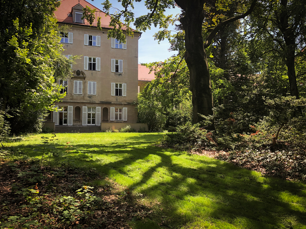 Das Hauptgebäude des Verlags C.H.Beck, umgeben von einem grünen, baumbestandenen Garten an einem sonnigen Tag.