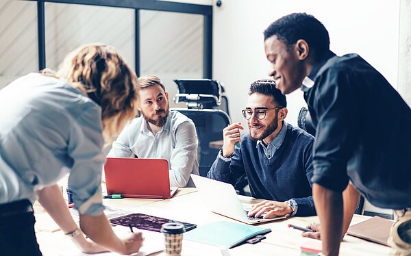 Ein Team von vier Personen sitzt und steht um einen Konferenztisch, diskutiert und arbeitet zusammen. Einige Teilnehmer haben Laptops vor sich, während eine Person Notizen auf einem Tablet macht. Die Stimmung ist konzentriert und produktiv.