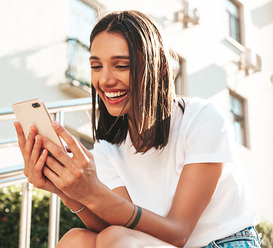 Frau in einem weißen T-Shirt lächelt, während sie ihr Smartphone im Freien benutzt.